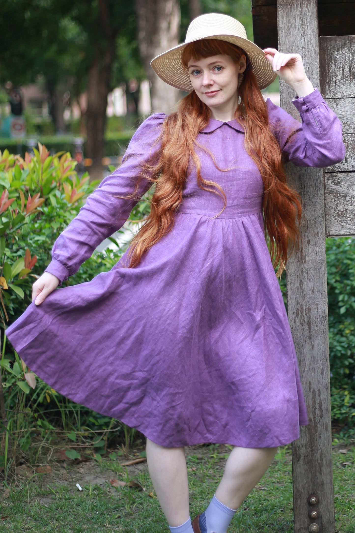 Woman in a purple dress and white hat standing in a garden.